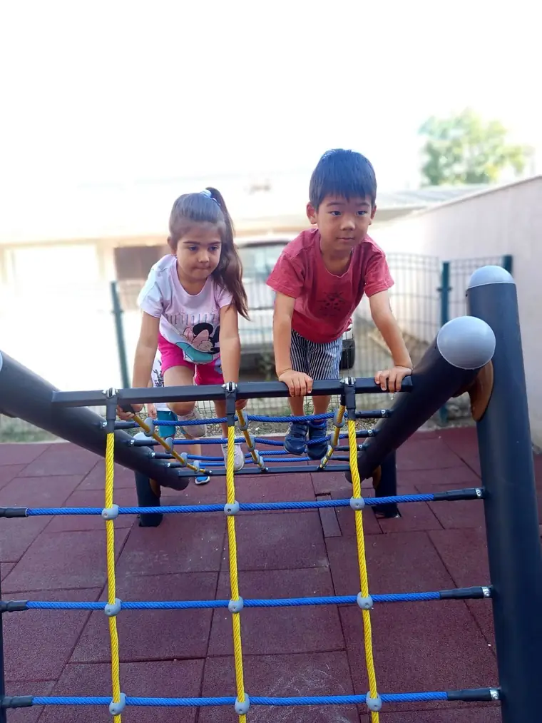 Devojčica i dečak se penju uz mrežu od plave i žute užadi na igralištu