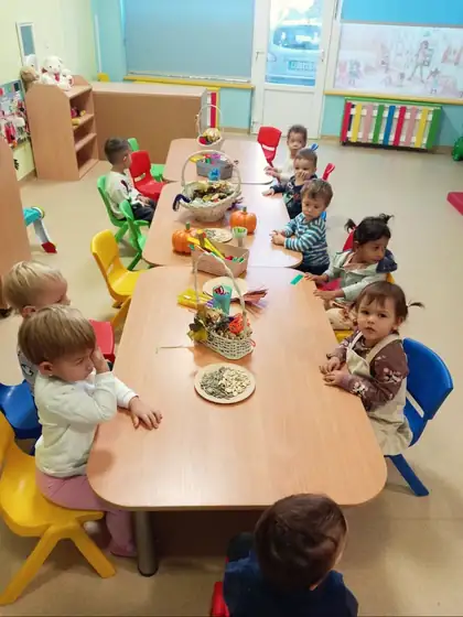 Mališani iz jaslene grupe za niskim stolom sa korpama jesenjeg lišća i plodova