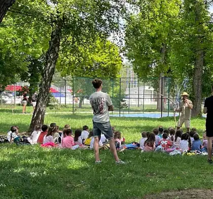 Deca sede na travi u parku, a ispred njih mladić vodi igru na otvorenom